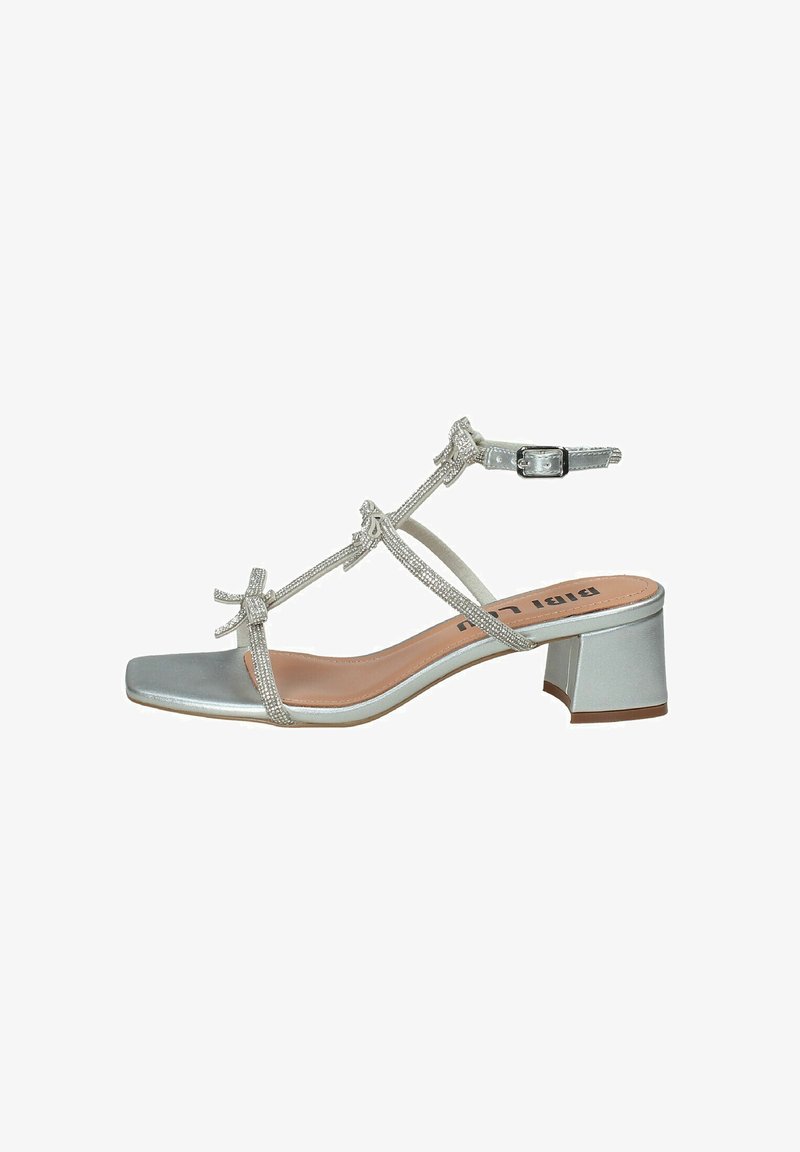 Sandalo argentato con cinturini e punta quadrata, con tacco basso a blocco. Caratterizzato da fiocchi decorativi e chiusura con fibbia. Materiale sintetico liscio.