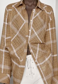 Bruine linnen shirt met een wit ruitpatroon, knoopsluiting aan de voorkant, lange mouwen, relaxte pasvorm. Witte vetersluiting details op de broek hieronder.