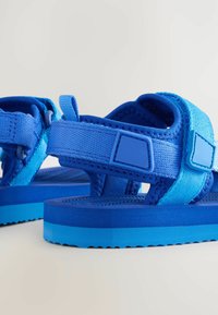 Blå sandaler med justerbara tygremmar, texturerade innersulor och en hållbar gummisula. Har kontrasterande nyanser av blått genom hela.