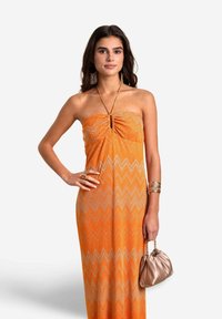 Abito lungo senza spalline di colore arancione con un motivo a zigzag, in tessuto leggero e con dettagli arricciati sul corpetto; abbinato a una piccola borsa dorata.
