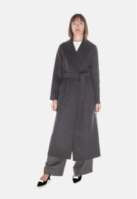 Cappotto lungo in misto lana di colore grigio scuro con design avvolgente, ampi revers e cintura da annodare. Il modello indossa pantaloni grigi e scarpe nere con dettagli argentati.