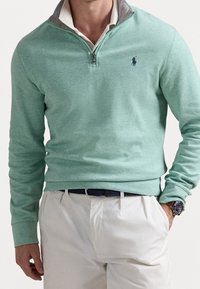Pullover verde chiaro con design a zip quarti e dettagli grigi nel colletto. Tessuto morbido con maniche lunghe. Indossato con pantaloni bianchi e un orologio.