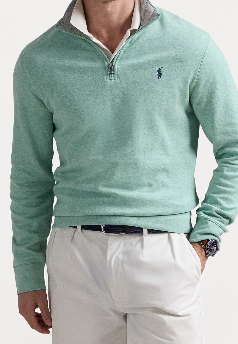 Pullover verde chiaro con design a zip quarti e dettagli grigi nel colletto. Tessuto morbido con maniche lunghe. Indossato con pantaloni bianchi e un orologio.