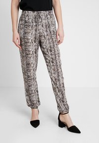 Kvinna iförd joggerbyxor med brunt och beige ormtryck i kombination med svarta klackar med ankelrem på en vit bakgrund.