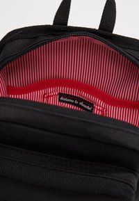 Wnętrze czarnego plecaka z tkaniny, z czerwono-białą paskowaną podszewką, etykietą z napisem „Welcome to Herschel” oraz gładką teksturą.