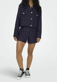 Femme portant une veste boutonnée texturée bleu marine foncé avec un short assorti et des baskets montantes noires à lacets et semelles blanches.