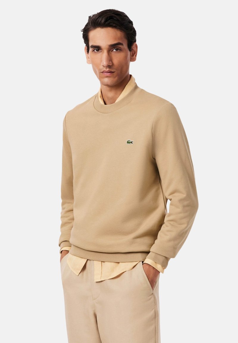 Sudadera beige con cuello alto y puños acanalados, con un pequeño logo verde. Se lleva sobre una camisa amarilla clara con cuello.