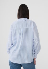 Donna con capelli lunghi e scuri che indossa una camicia a righe verticali bianca e azzurra a maniche lunghe e un paio di jeans blu, di spalle.
