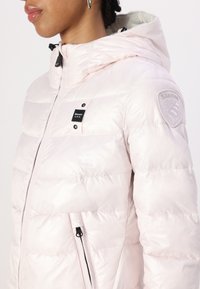 Giacca imbottita rosa chiaro con cappuccio, caratterizzata da una trapuntatura orizzontale, accessori neri, una patch con logo sulla manica e due tasche laterali con cerniera.