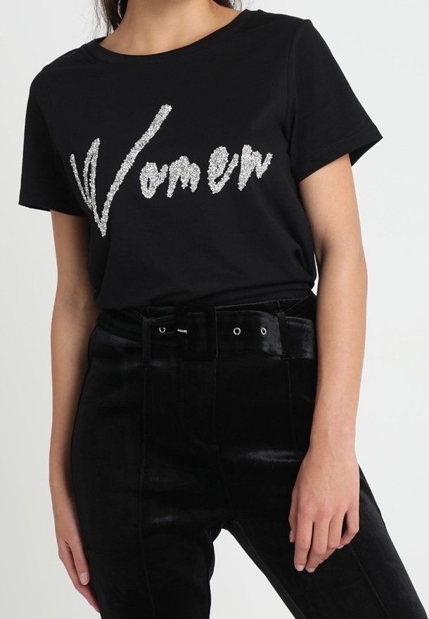 Mujer vistiendo una camiseta negra de manga corta con la palabra "Women" en texto de purpurina plateada, combinada con pantalones de terciopelo negros de talle alto y cinturón.