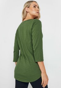 Maglia a maniche lunghe di colore verde oliva, con scollatura a barchetta e orlo arrotondato. Realizzata in un tessuto morbido e strutturato con maniche a 3/4.