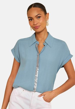 Femeie purtând o bluză albastru deschis cu mâneci scurte, cu nasturi decorați cu paiete și pantaloni albi, pozând cu mâna în buzunar.