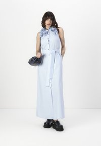 Απαλή μπλε, μακριά φόρεμα πουκάμισο με μπροστινό κουμπί και ζωνάκι στη μέση; διαθέτει λουλούδια από ύφασμα. Το μοντέλο κρατά μια μαύρη τσάντα.