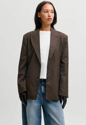 Femme portant un blazer marron à fines rayures sur une chemise blanche, un jean bleu et des gants noirs, regardant légèrement vers sa gauche sur un fond uni.