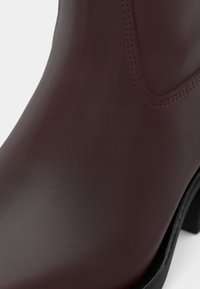 Primer plano de una bota de ankle de cuero marrón oscuro, suave, con panel elástico lateral y suela y tacón negros.