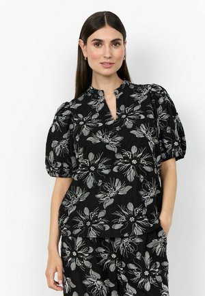 Vrouw met lang donker haar die een zwarte top en broek met grote witte bloemmotieven draagt, staande met één hand in de zak tegen een witte achtergrond.