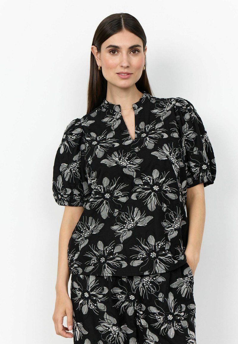 Femme aux longs cheveux foncés portant un haut noir et un pantalon avec de grands motifs floraux blancs, debout avec une main dans la poche sur fond blanc.