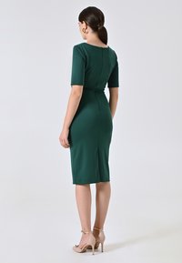 Grünes, tailliertes Kleid mit kurzen Ärmeln, einem Reißverschluss auf der Rückseite und knielangem Saum. Glatte Textur, betonte Taille, kombiniert mit hautfarbenen High Heels.