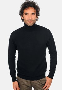 Schwarzer Rollkragenpullover, gestrickte Textur, lange Ärmel, figurnahes Design, gerippte Bündchen und Saum, kombiniert mit braunen Hosen.