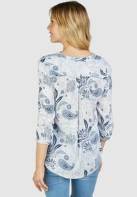 Weiße Bluse mit blauen Paisleymustern, Dreiviertelärmeln und rundem Ausschnitt. Leichter Stoff mit Falten-Detail auf der Rückseite.