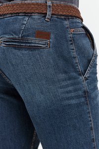 Mörk indigo denim jeans med ett flätat brunt bälte. Har en logotyp i form av en lapp på bakfickan och kontrasterande sömmar vid sömmarna.