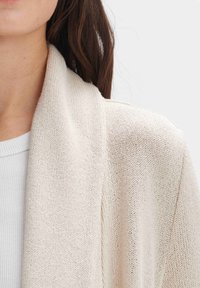 Helle beigefarbene Strickjacke mit strukturiertem, offenem Strickdesign und breiten Revers. Getragen über einem weißen, gerippten Oberteil. Weiches, atmungsaktives Material.