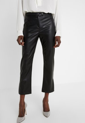 Vrouw draagt zwarte leren cropped broek, witte blouse met lange mouwen en zilveren hoge hakken, staand tegen een witte achtergrond.