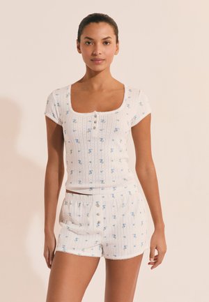 Donna che indossa un top a maniche corte a coste bianco e pantaloncini abbinati con una piccola stampa floreale blu e dettagli con bottoni.