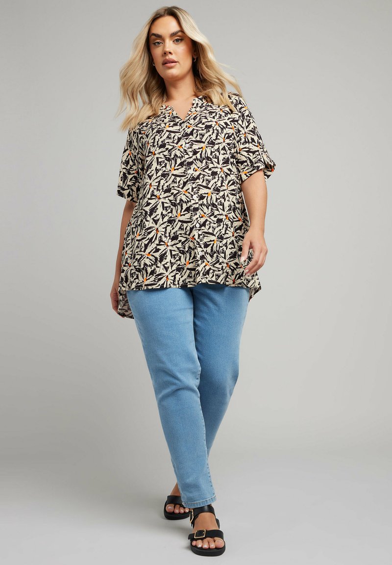 Camicia leggera a maniche corte di colore crema con motivo tropicale di foglie nere e arancioni, abbinata a jeans blu chiaro e sandali neri.