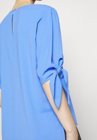 Femme portant un chemisier bleu aux manches trois-quarts nouées en nœuds près des coudes, vue de dos sur un fond uni.