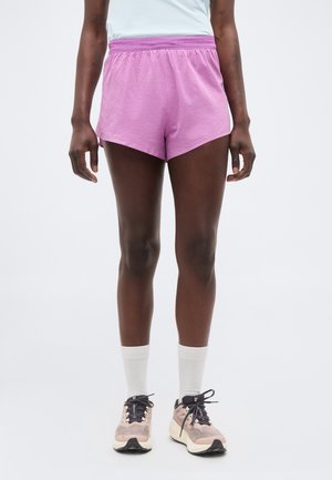 Πόδια ενός ατόμου που φορά ροζ αθλητικά σορτς, λευκές κάλτσες μέχρι τον αστράγαλο και ροζ-μαύρα παπούτσια για τρέξιμο, και στέκεται μπροστά σε απλό φόντο.