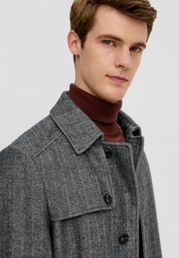 Grau karierten Mantel mit einem spitzen Kragen, der an Wolle erinnert, mit schwarzen Knöpfen und über einem bordeauxfarbenen Rollkragenpullover getragen.