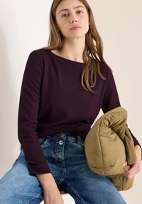 Ribbineulinen, pitkähihainen tummanvioletti paita yhdistetty sinisiin farkkuihin. Pehmeä, khakinvärinen untuvatakki lepää sylissä. Sileä ihonsävy ja suora hius.