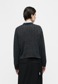 Άτομο με κοντά σκούρα μαλλιά, που φοράει σκούνο πουλόβερ με πλεκτό σχέδιο και μαύρη φούστα, εμφανιζόμενο από πίσω, με απλό λευκό φόντο.