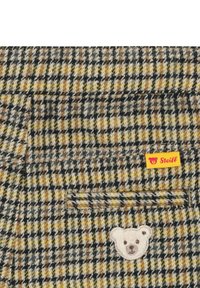 Tela de jaquard en marrón y amarillo con una textura especial, que presenta un parche con el logo de un oso y una etiqueta amarilla "Steiff" en un bolsillo.