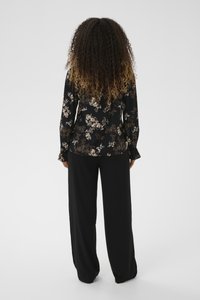 Ženska s dolgimi kodrastimi lasmi, oblečena v črno cvetlično majico z dolgimi rokavi in ohlapne črne hlače, stoji obrnjena proč proti nevtralnemu ozadju.