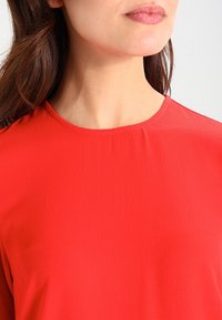 Rode, kort mouwtop met ronde hals. Gemaakt van gladde, lichte stof met een subtiele textuur. Plooidetail bij de hals.