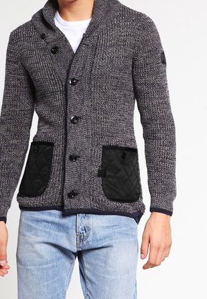 Cardigan grigio lavorato a maglia con tasche applicate nere trapuntate, chiusura con bottoni sul davanti, indossato sopra una camicia bianca e jeans azzurri chiari.