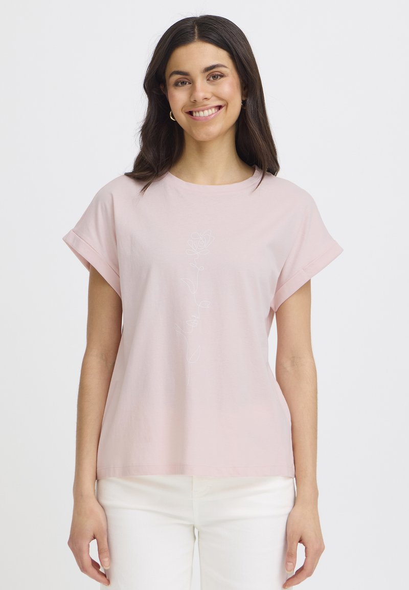 Junge Frau lächelnd, trägt ein hellrosa Kurzarm-Top mit Rosendesign und weiße Hose, steht vor einem schlichten weißen Hintergrund.