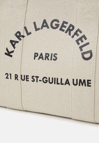 Beige canvas shoppingväska med fet svart text: "KARL LAGERFELD PARIS" och "21 RUE ST-GUILLAUME." Enkel, strukturerad design.