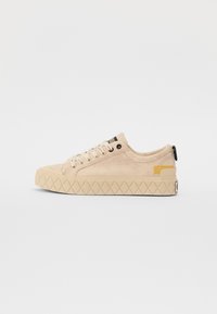 Beige sneaker with a textured fabric upper, diagonal stitching, and a rubber sole featuring a diamond pattern. Yellow accent on heel.