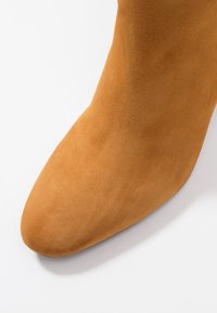 Primer plano de un zapato de ante color beige con puntera redondeada sobre un fondo blanco.