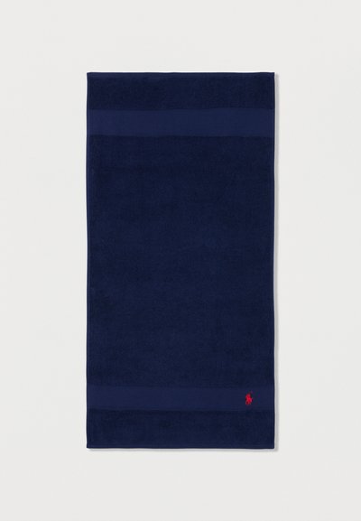 Σκούρα μπλε ορθογώνια πετσέτα με δύο οριζόντιες υφασμένες λωρίδες, με ένα μικρό κόκκινο κεντημένο λογότυπο παίκτη πόλο κοντά στην κάτω δεξιά γωνία.