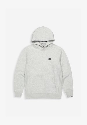 Man in een bijpassende lichtgrijze hoodie en joggingbroek, staand met één hand in de zak van de hoodie tegen een effen achtergrond.