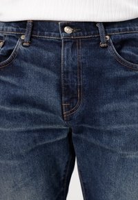 Jeans de denim azul oscuro con un corte clásico. Las características incluyen un botón metálico, costuras naranjas en contraste y bolsillos frontales. Textura suave.