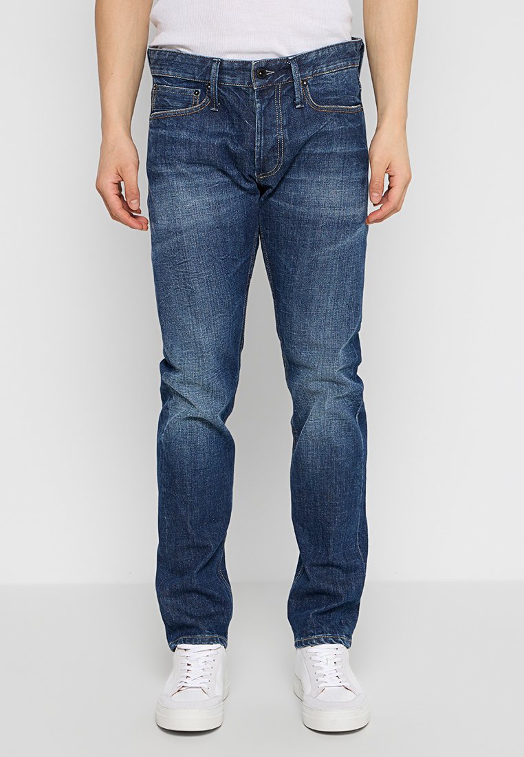 Donkerblauwe denim jeans met een rechte pasvorm, voorzien van vijf zakken, subtiele whiskering en contrasterende stiksels, gecombineerd met witte sneakers.