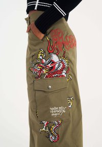 Olivgrüne Cargo-Hosen mit roter und gelber Drachenstickerei, ausgestattet mit einer großen Seitentasche und silbernen Hardware-Details. Handgefertigtes Logo sichtbar.