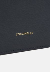 Proizvod od plave teksturirane kože s zlatnim logotipom "COCCINELLE", šivenim rubovima i glatkom, mat površinom.