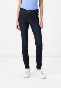 Pantalones vaqueros skinny azul oscuro de denim, con un diseño clásico de cinco bolsillos y costuras doradas en contraste, combinados con zapatillas blancas.