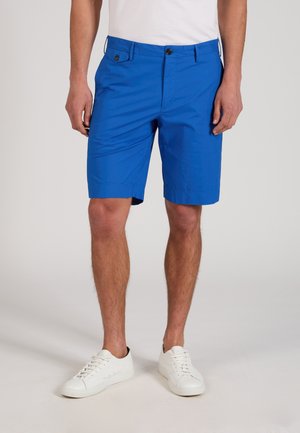 Hombre con pantalones cortos azules brillantes hasta la rodilla, zapatillas blancas y camisa blanca, de pie frente a un fondo liso.